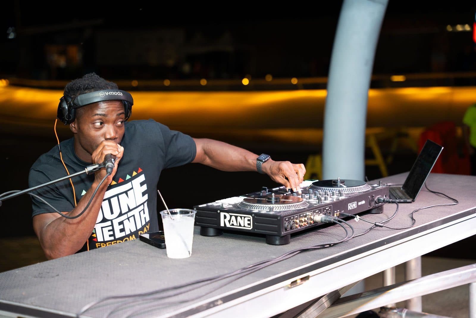 A DJ in headphones speaks into a microphone while adjusting a controller at an outdoor night setup with warm lights behind
