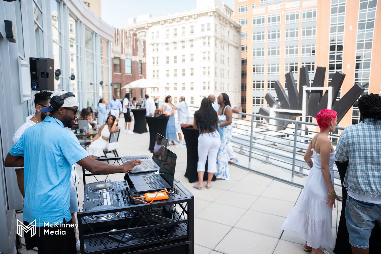 A DJ in a light blue shirt at a controller on a sunny rooftop terrace while guests dressed in white mingle nearby