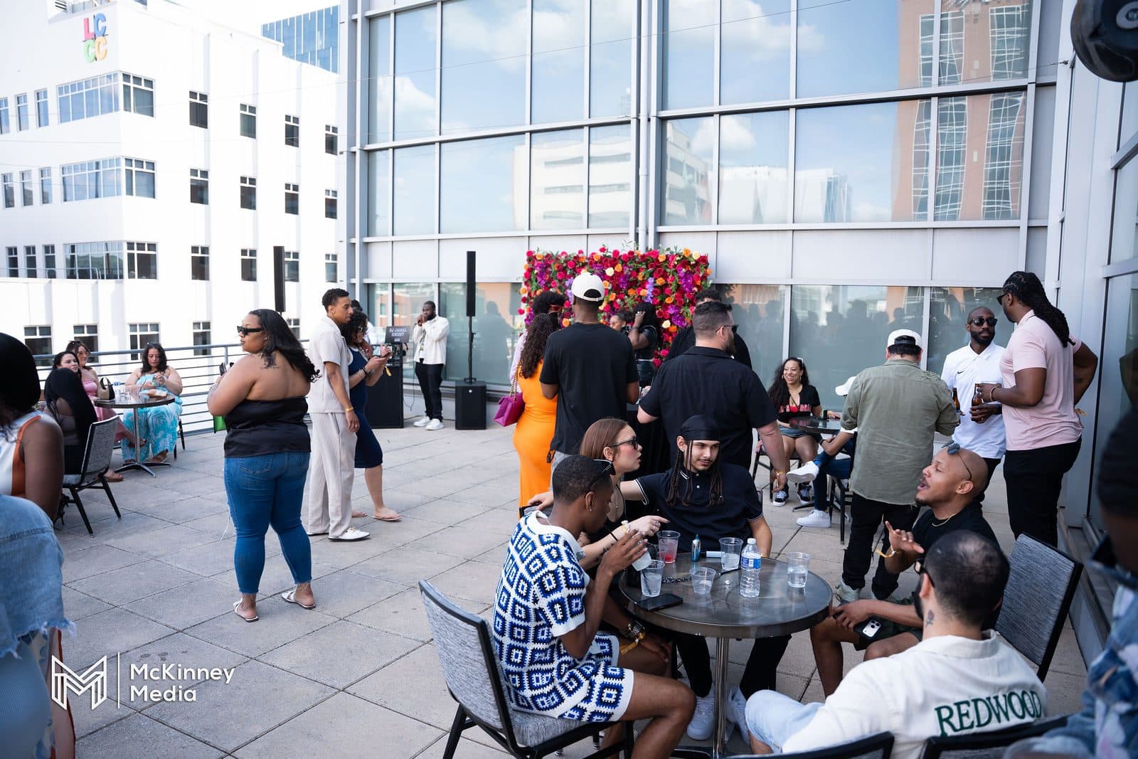 Guests at tables and standing on a rooftop terrace surrounded by glass buildings, with a flower wall in the background