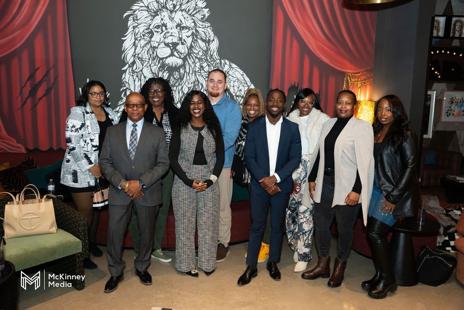 About a dozen professionally dressed attendees posing together in a lounge with red curtains and a large lion mural