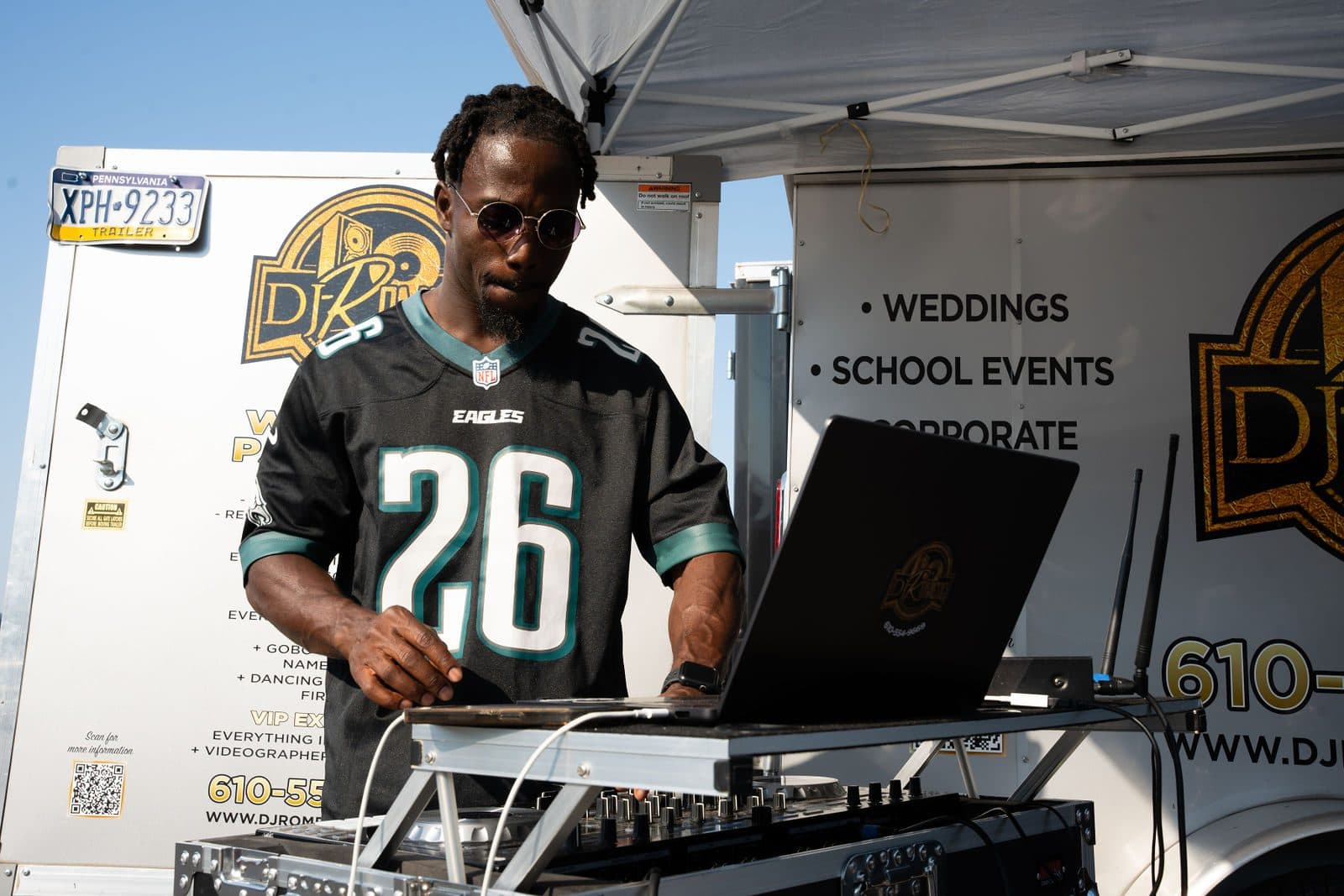 A DJ in sunglasses and an Eagles jersey works a laptop and controller in front of a branded trailer under a canopy