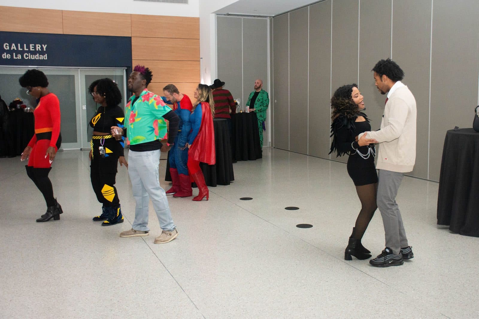Costumed guests, including two in caped superhero outfits, dancing on a bright open floor in a modern event hall
