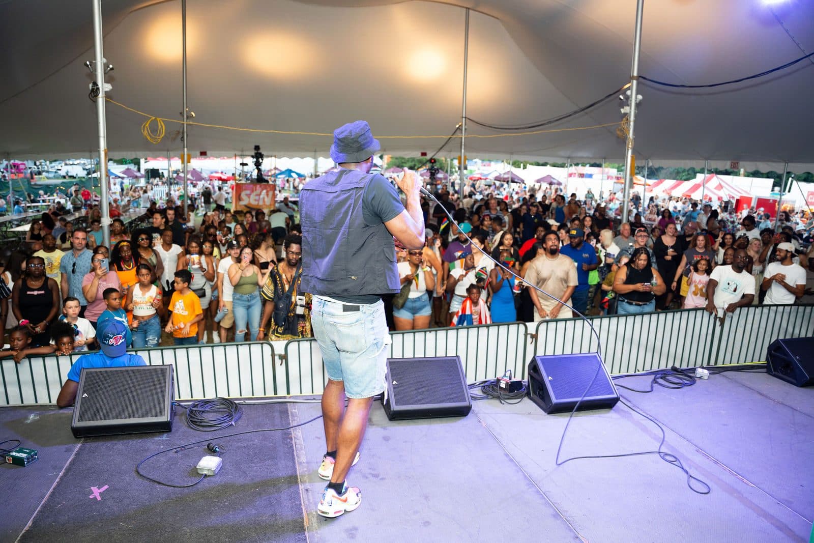 A performer with a microphone, seen from behind on a tent stage, faces a large crowd pressed against the barricade