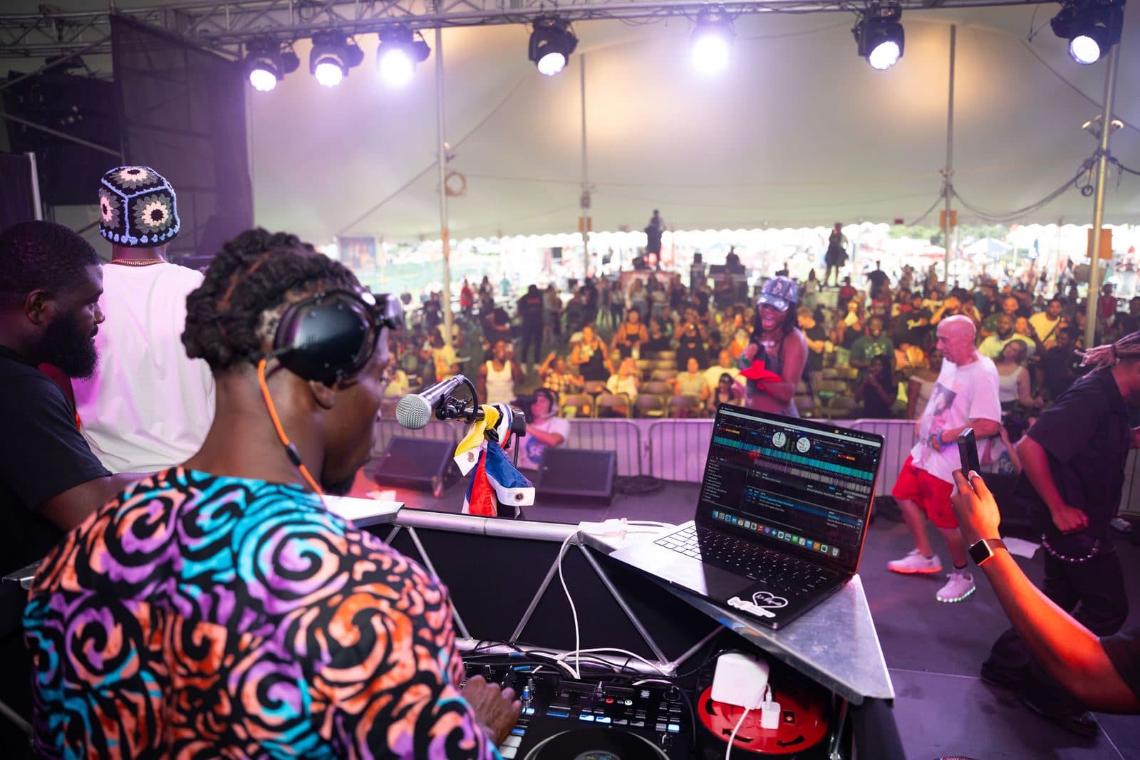 A DJ in headphones, seen from behind at the controller and laptop, with dancers on stage and a crowd under the tent