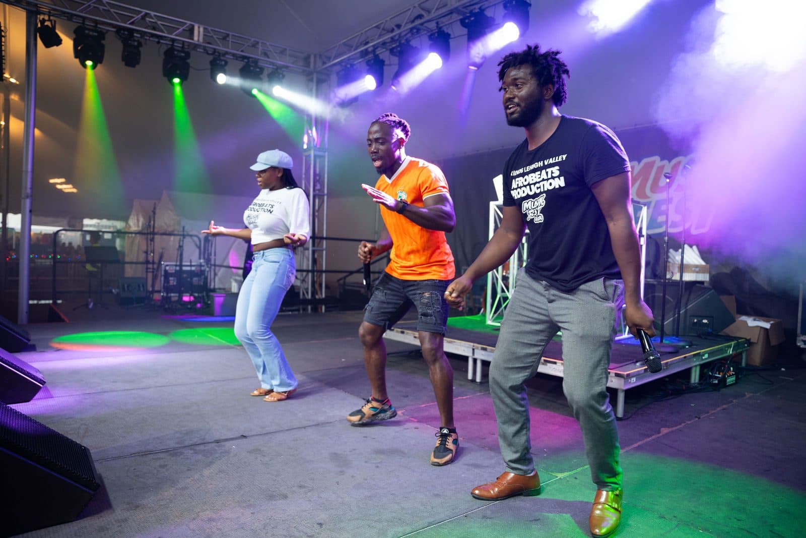 Three dancers performing on a festival stage lit with purple and green lights and haze