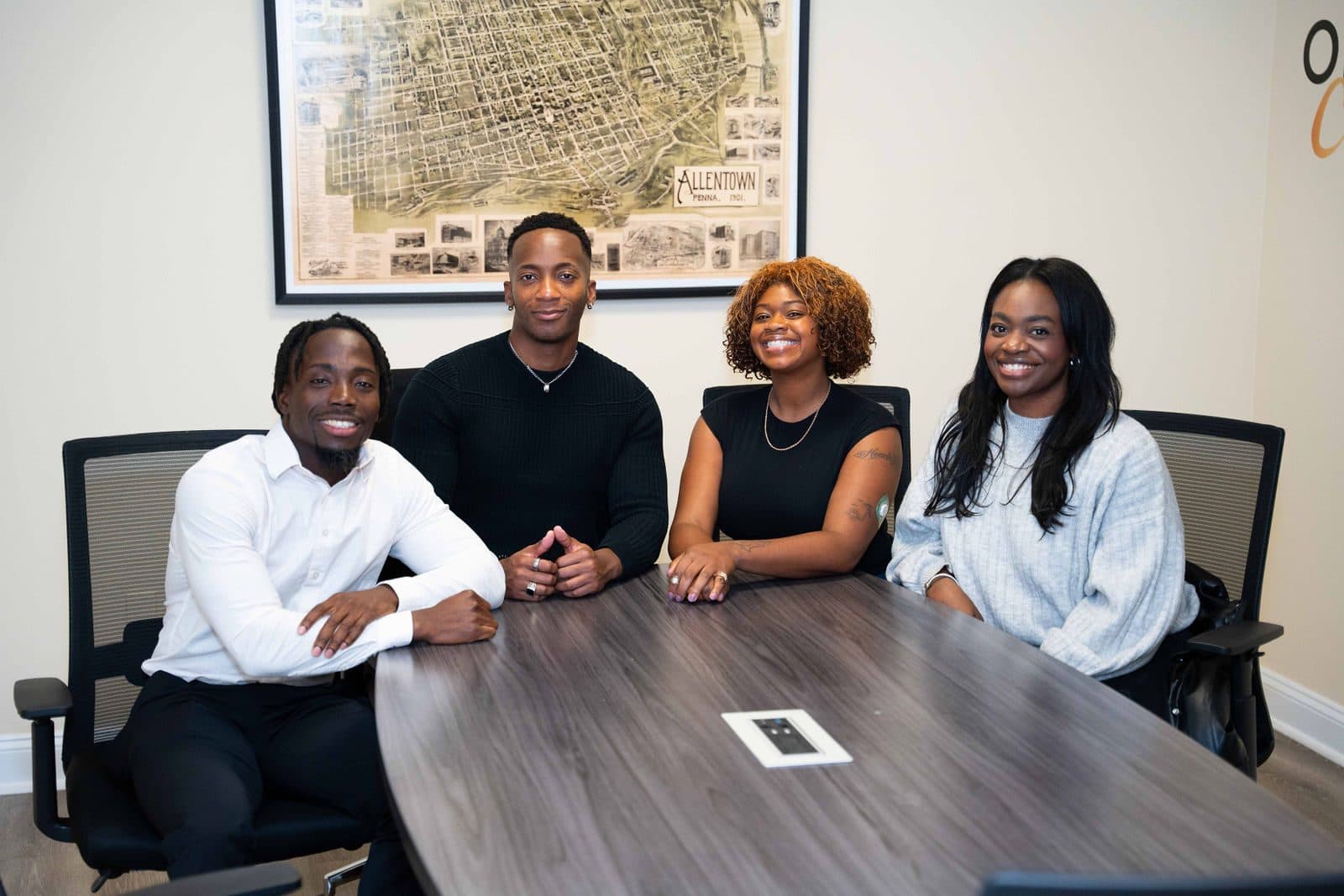 Four smiling people seated at a gray conference table in an office, with a framed map of Allentown on the wall