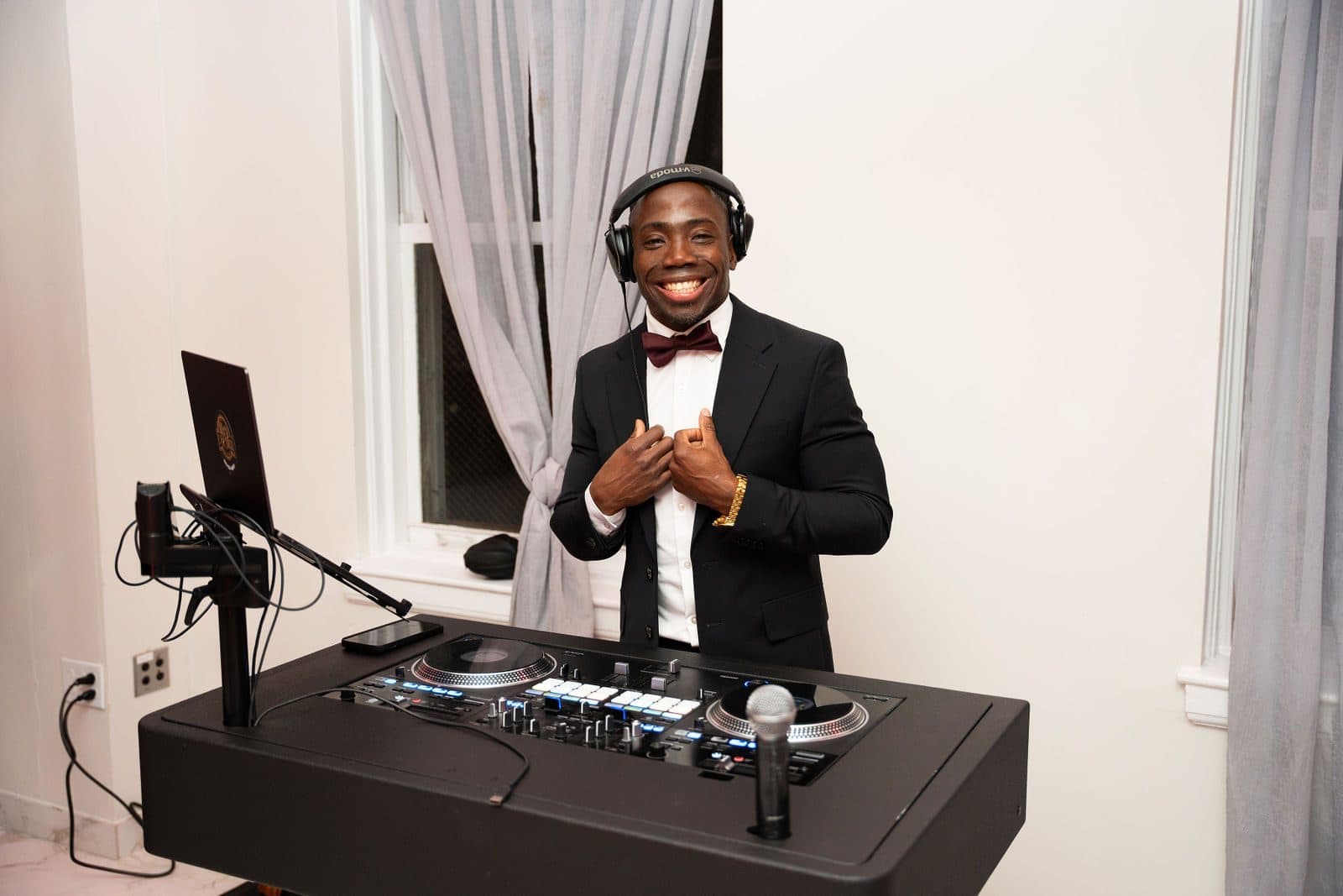 A smiling DJ in a tuxedo, bow tie and headphones behind a DJ console in a bright white room with sheer curtains