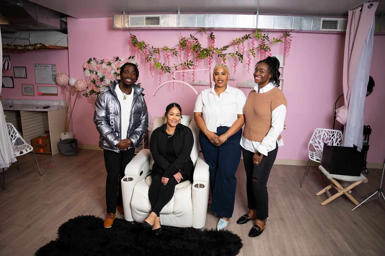 Four people posing in a pink studio, one seated in a white reclining chair, with flower garlands on the wall