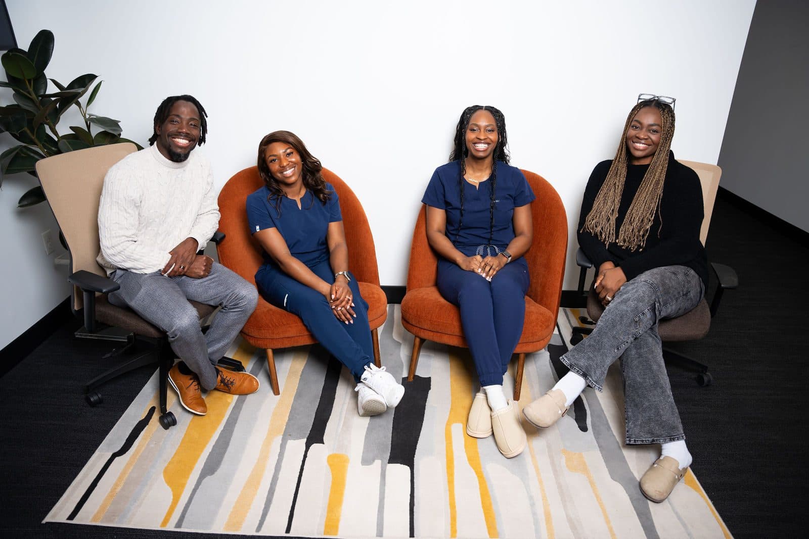 Four smiling people, two in navy scrubs, seated in a row of chairs on a striped rug in a bright white room