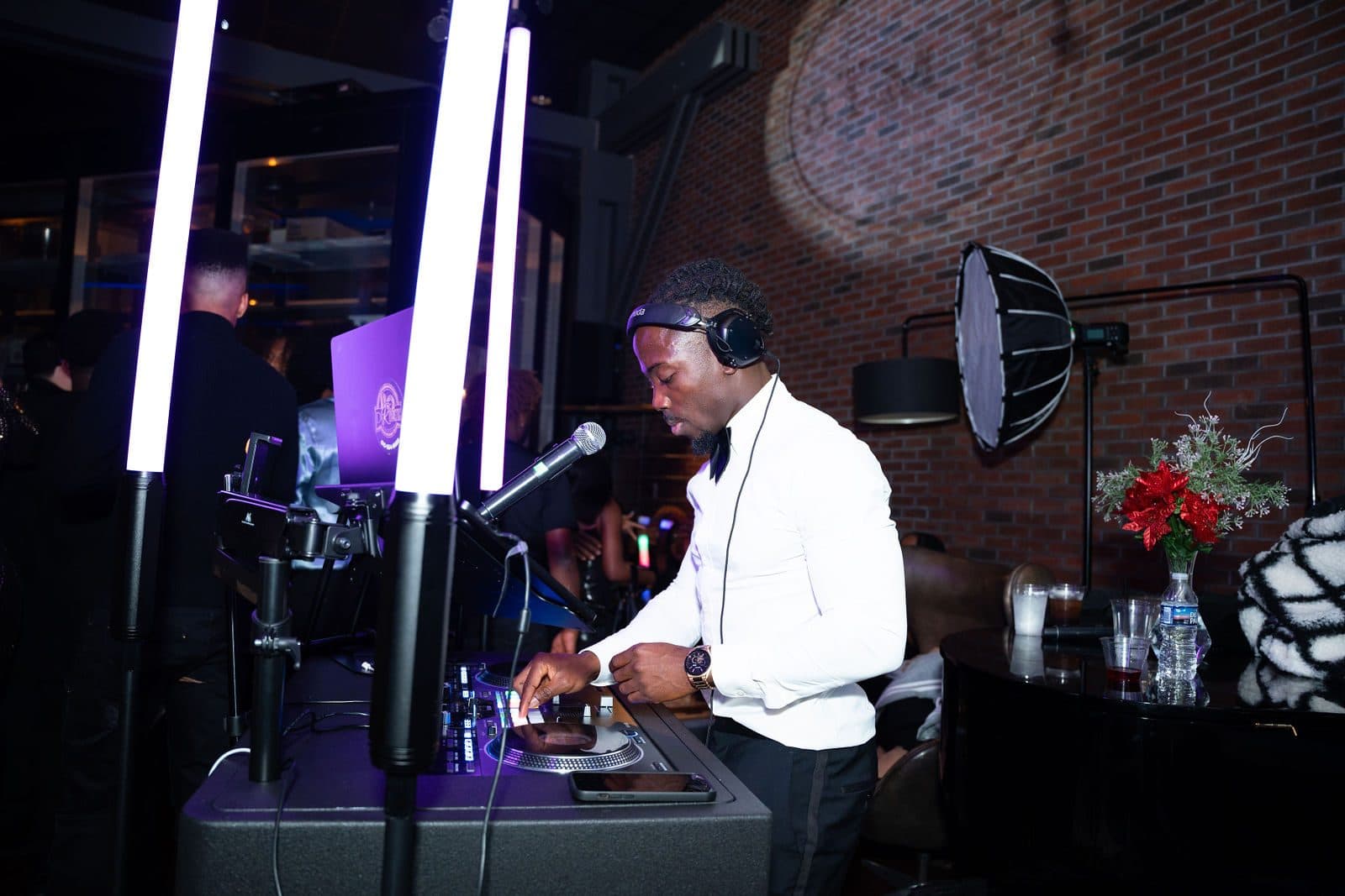 A DJ in a white shirt, bow tie and headphones at a console beside tall light bars and a red floral arrangement in a brick-walled bar