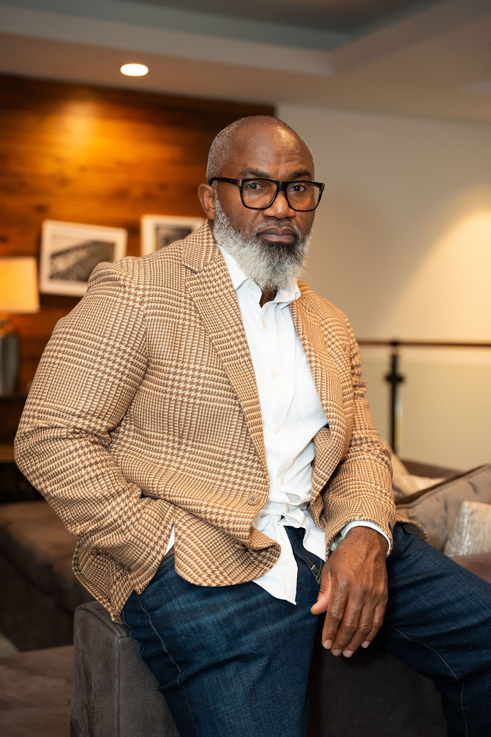 Portrait in a tan houndstooth blazer, seated on the arm of a sofa in a warmly lit lounge