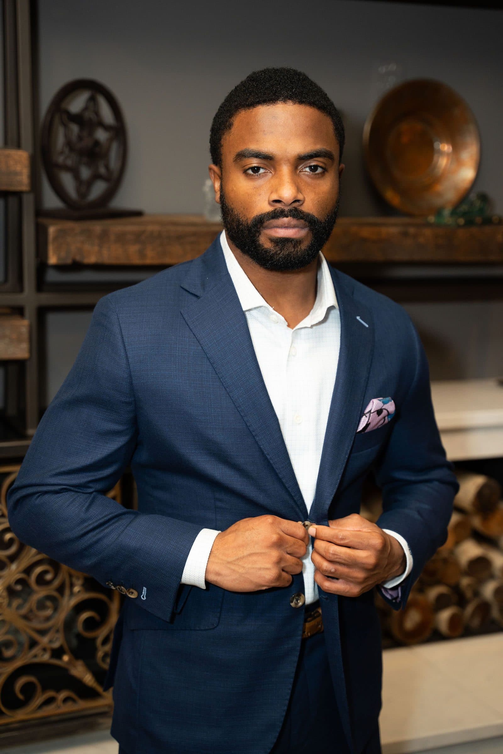 Portrait in a navy suit with a pocket square, buttoning the jacket in front of dark shelving