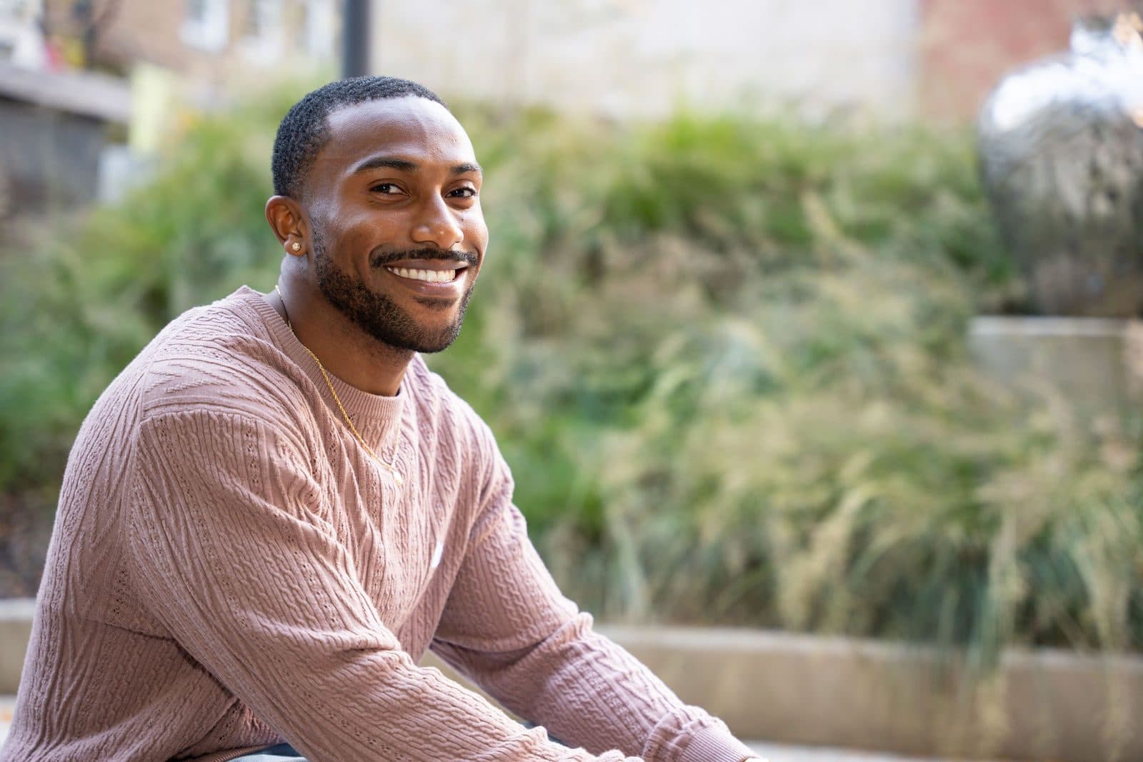 Smiling portrait in a pink knit sweater, seated outdoors in front of soft-focus plantings