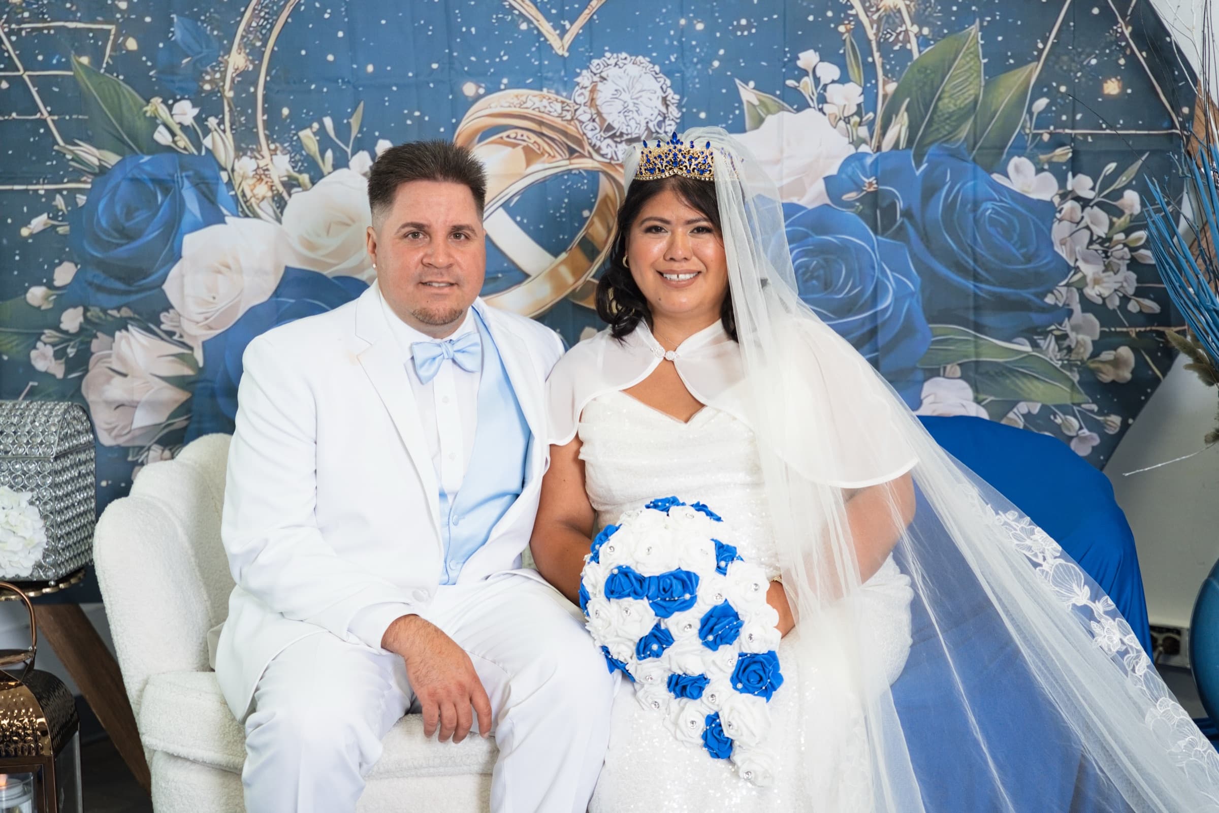 Maria and Jan Carlos seated together in white wedding attire against a blue-and-white floral backdrop
