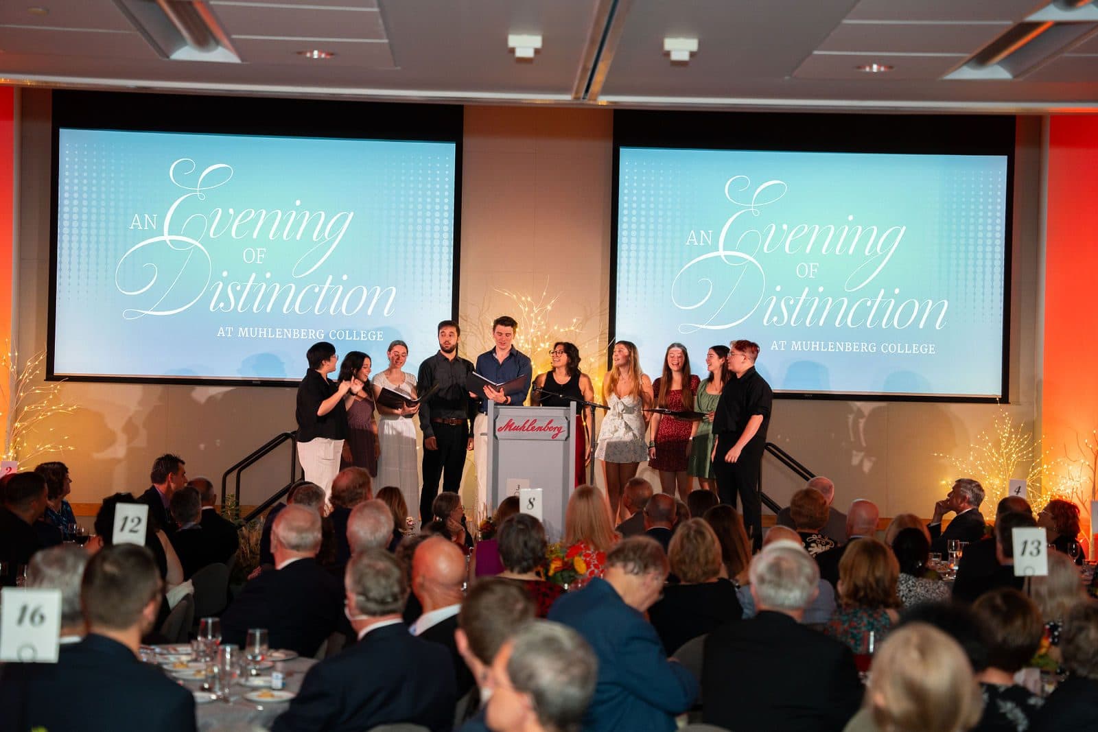Performers at the front of a dinner hall between two screens reading An Evening of Distinction at Muhlenberg College