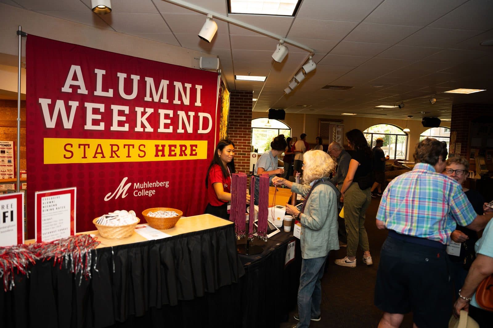 Check-in table beside a red banner reading Alumni Weekend Starts Here with the Muhlenberg College logo