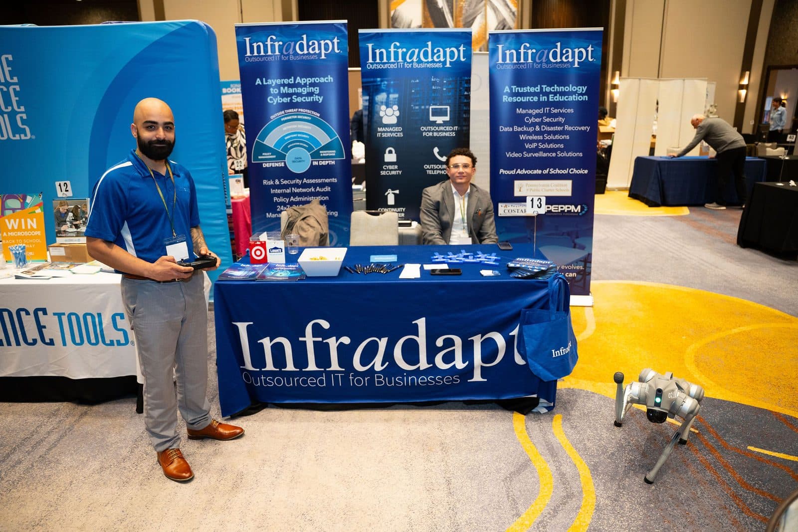 Exhibitor standing beside a branded booth with banners and a small robot on the floor in a conference exhibit hall