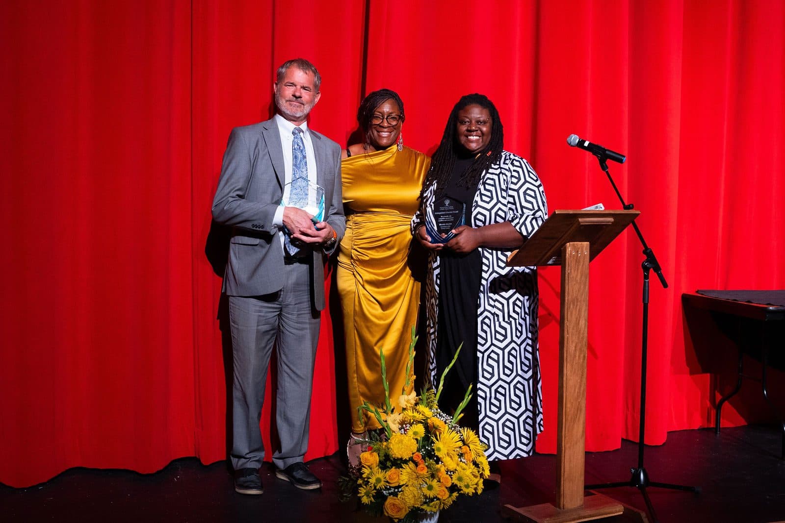 Three people on a stage with a red curtain holding glass awards, beside a podium and yellow flowers