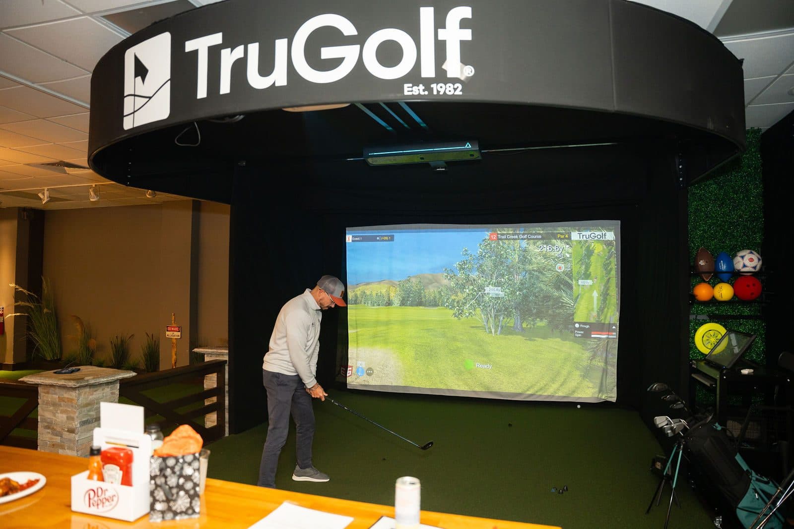 Guest swinging a club in an indoor golf simulator bay with a projected fairway on the screen