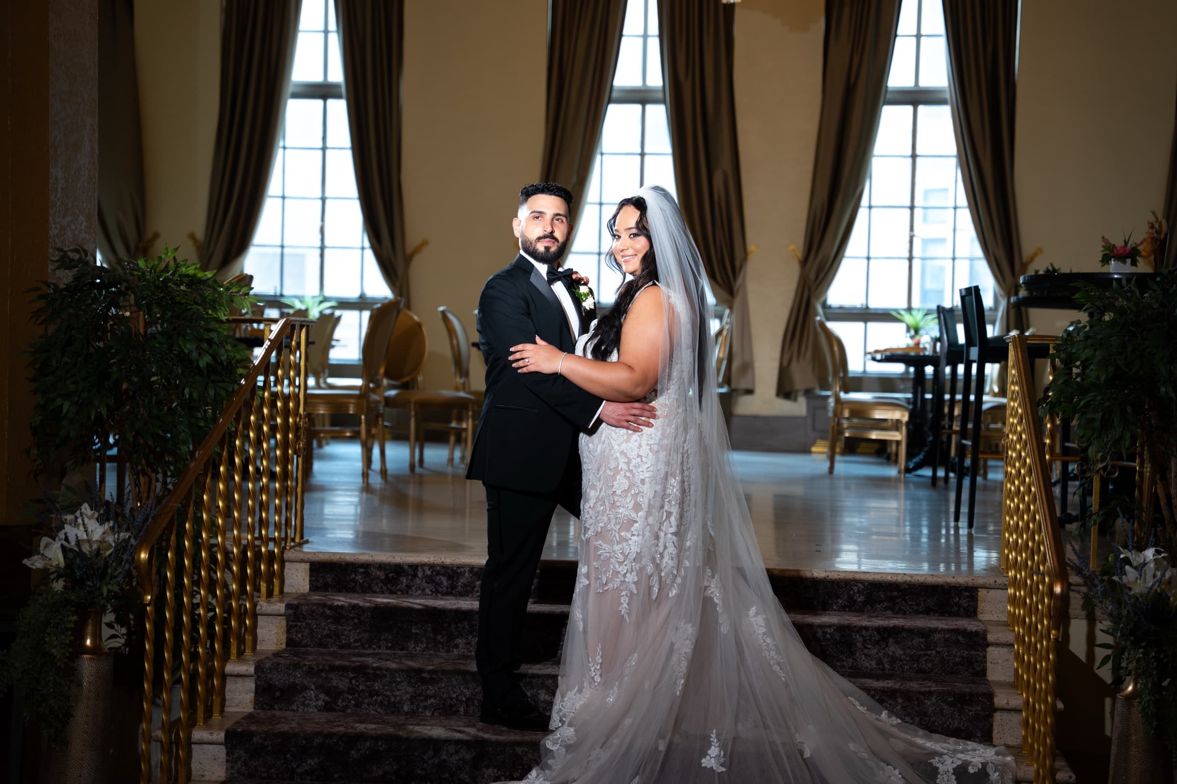Pauline y Sam abrazados en una escalera interior, con un vestido de encaje, ventanales altos y barandillas doradas