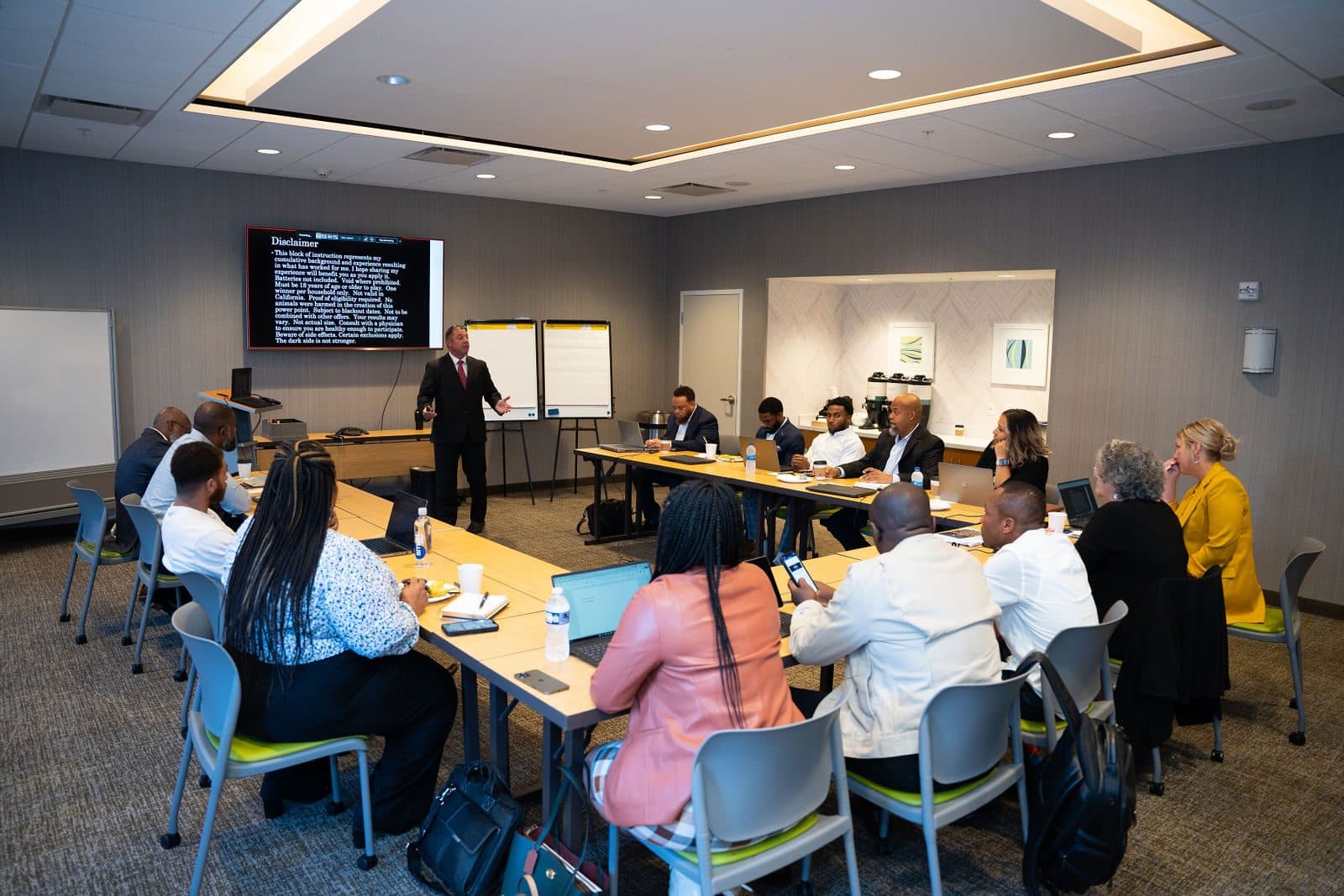 Presenter speaking beside a display screen to attendees seated at long tables in a bright conference room
