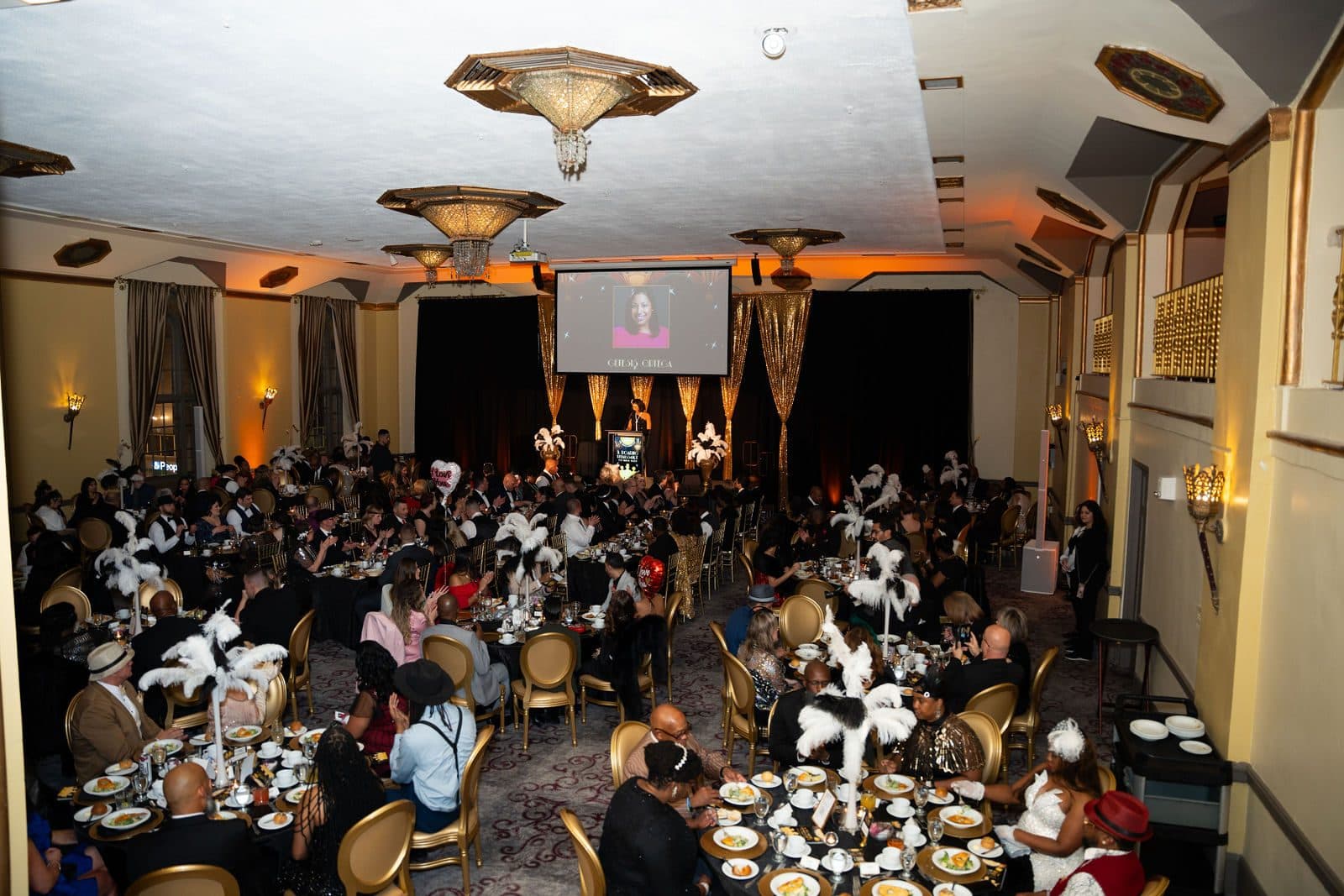 Wide view of a gala dinner in a ballroom with chandeliers and feathered centerpieces
