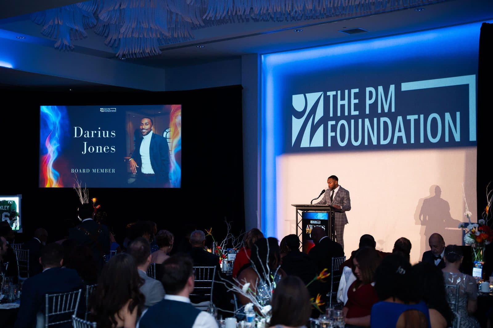 Speaker at a podium beside a large screen with The PM Foundation logo in a ballroom