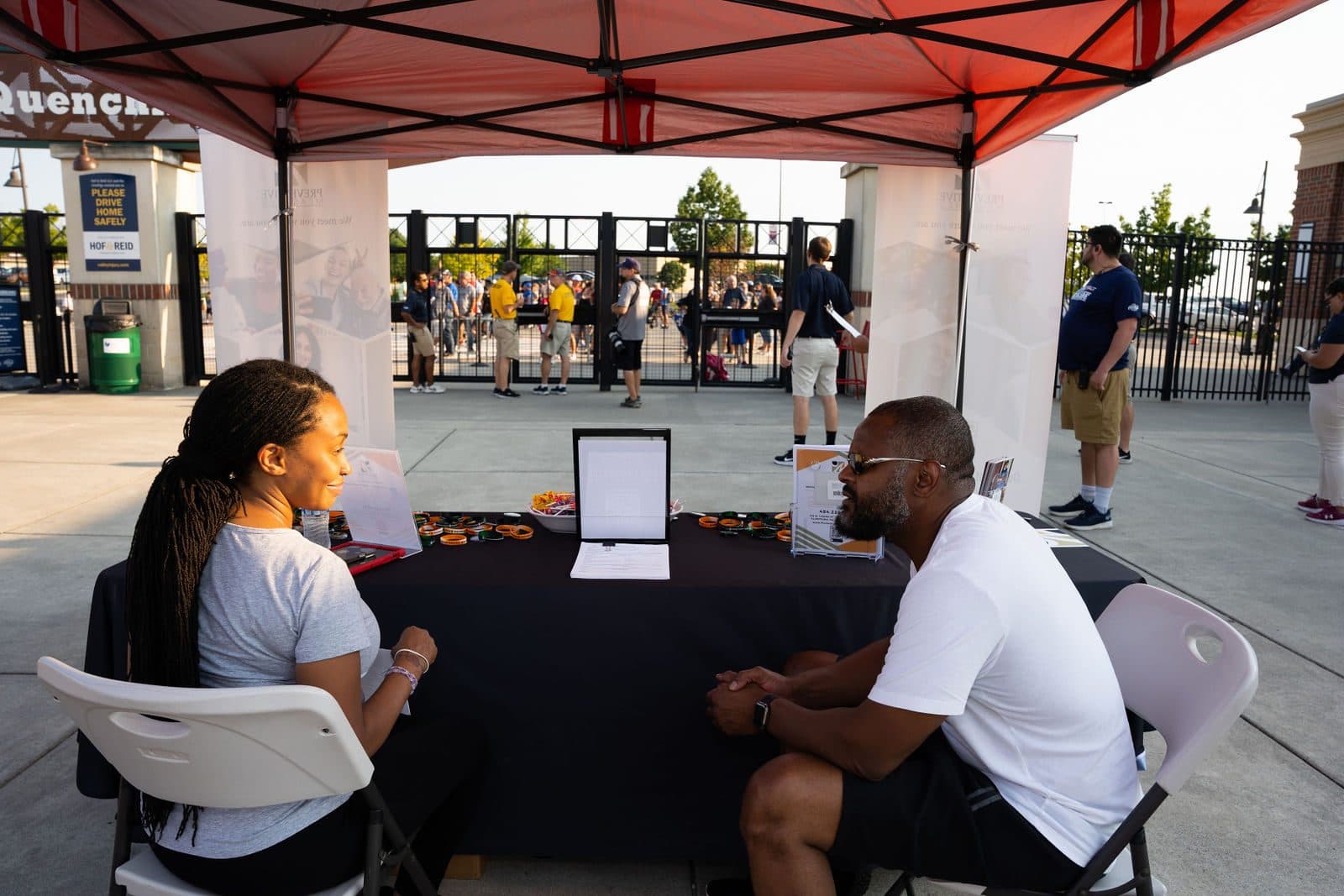 Two people seated at an information table under a red canopy at a ballpark entrance plaza