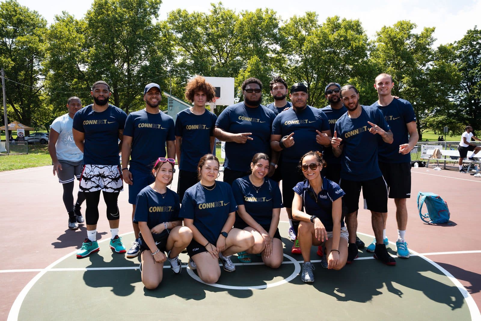 Team in matching navy shirts posing together on an outdoor basketball court surrounded by trees