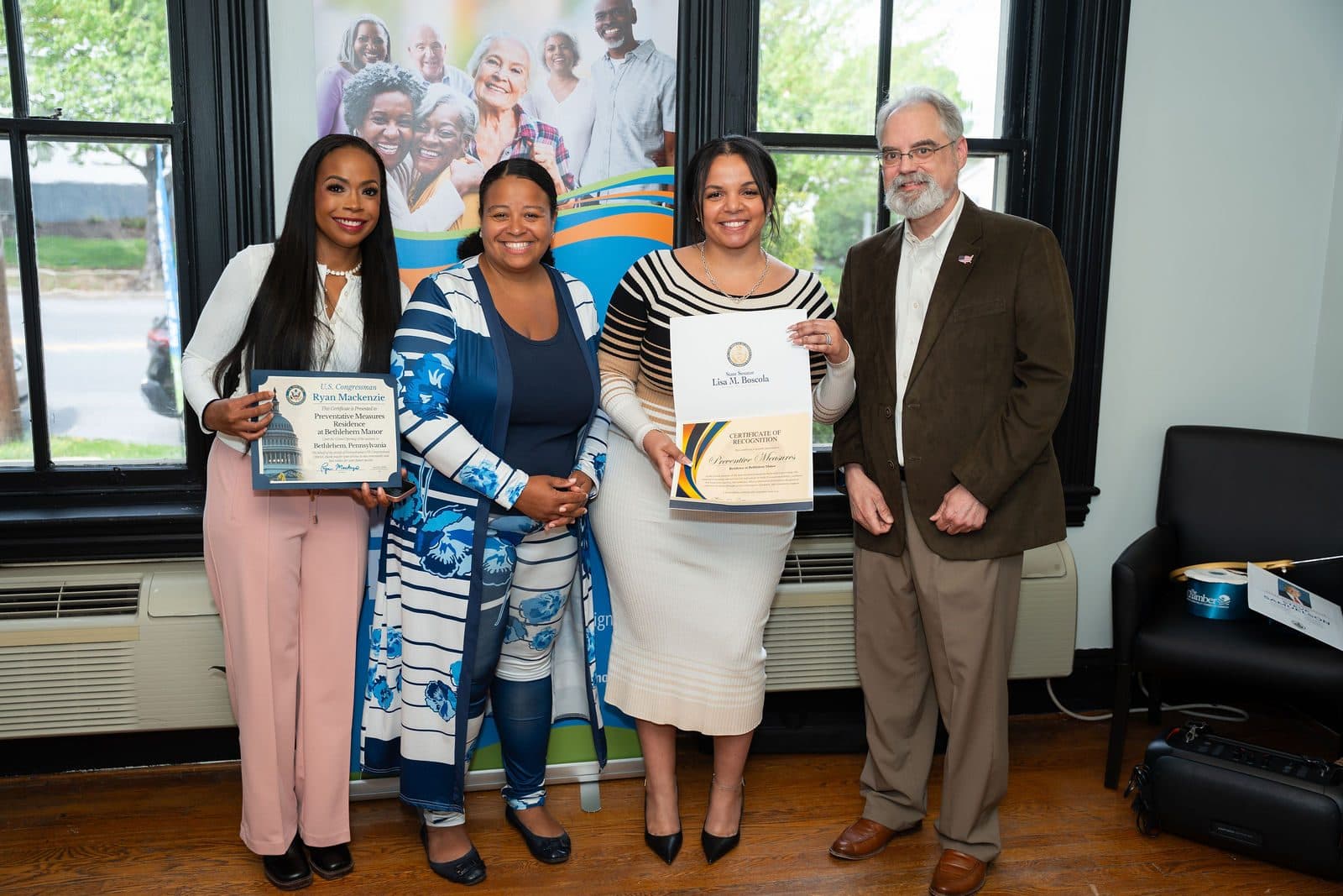 Four people posing with framed certificates in front of a PM Residence banner in a room with tall windows
