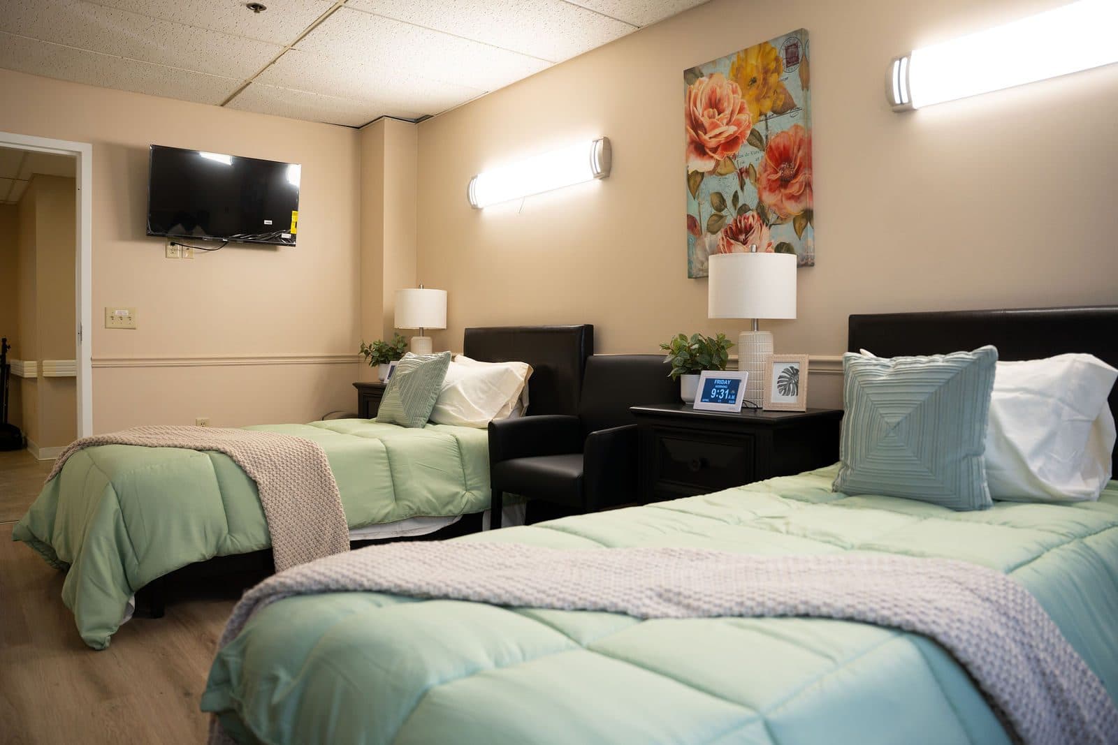 Furnished bedroom with two made beds in green bedding, bedside lamps, a wall-mounted TV, and floral artwork