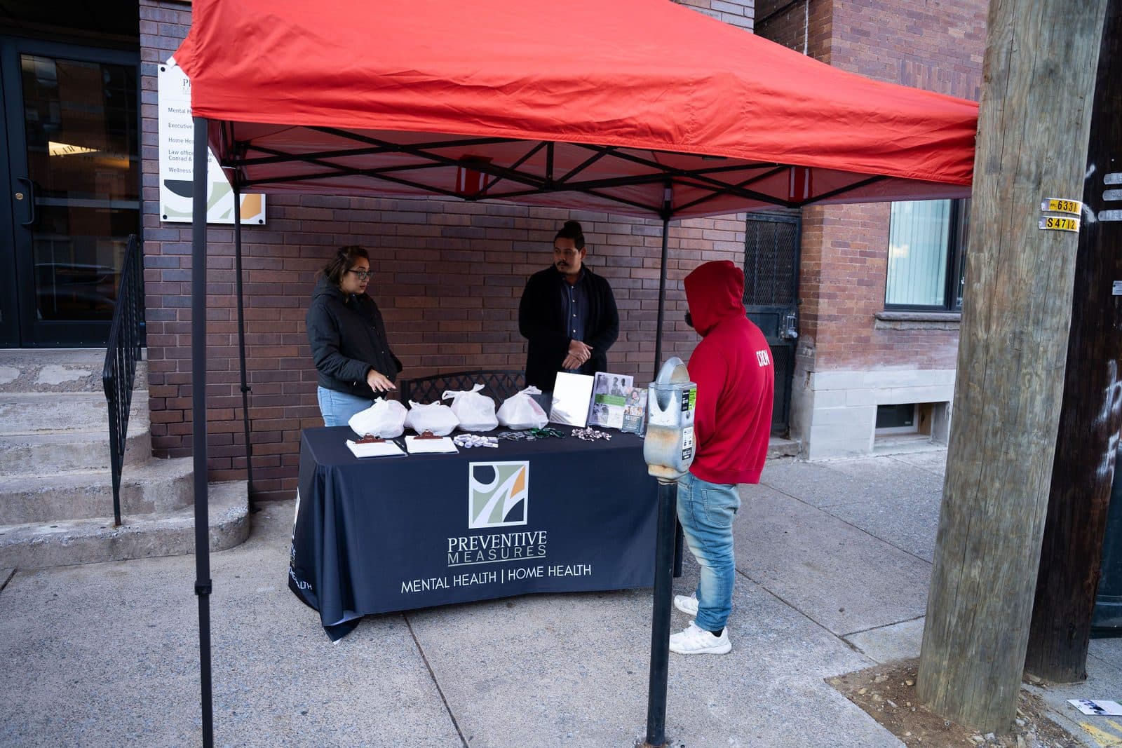 Volunteers at a Preventive Measures table under a red canopy on a city sidewalk, with bagged meals on the table