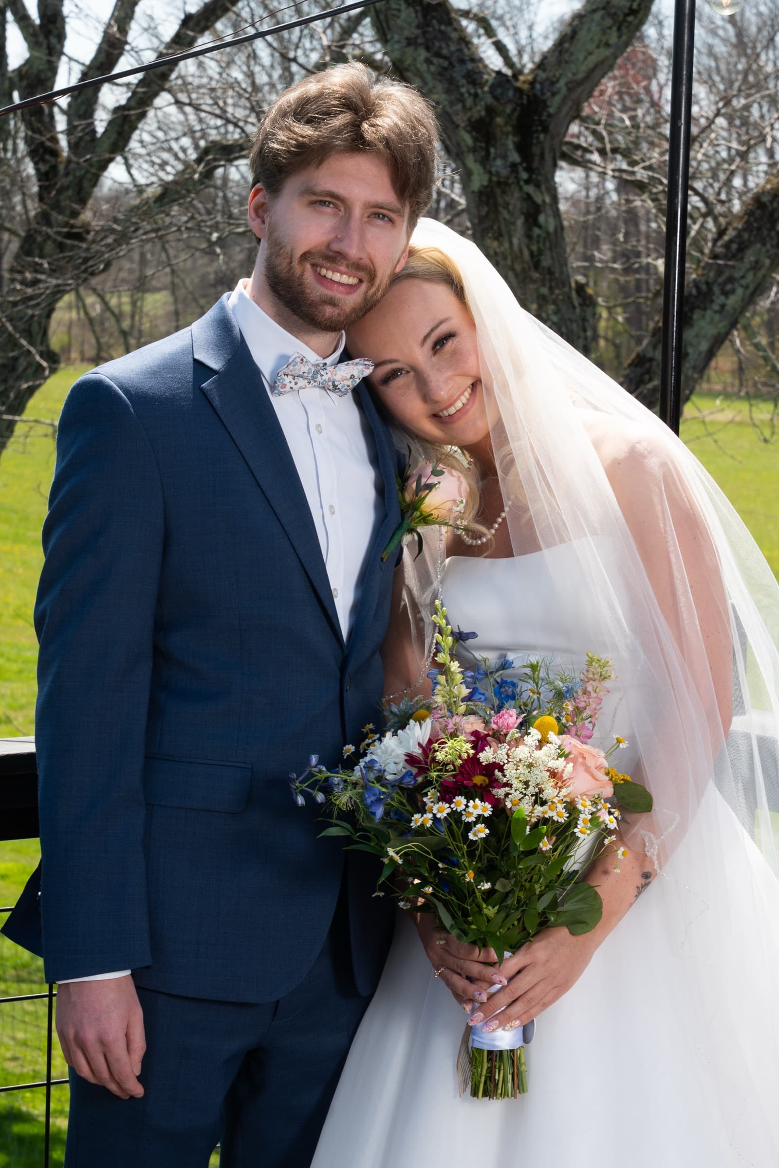 The Shimer couple leaning their heads together outdoors, holding a colorful bouquet beneath a white veil
