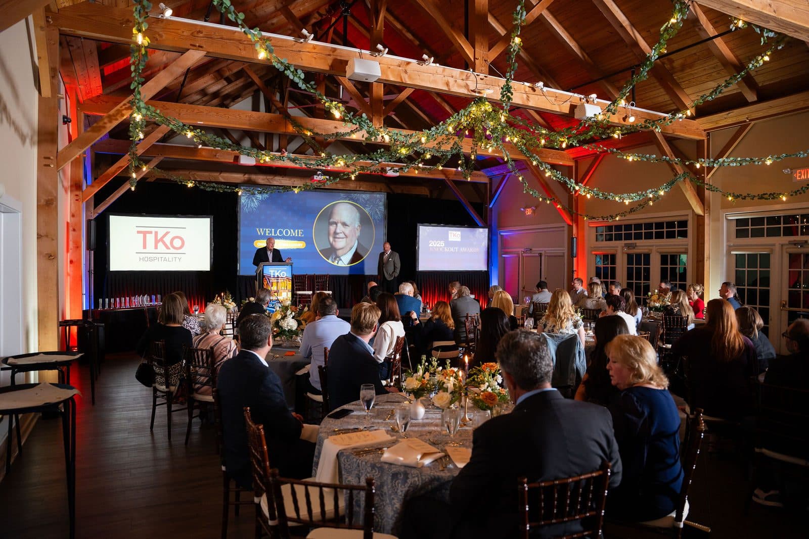 Guests at round dinner tables in a timber-beamed hall facing a stage with TKO screens