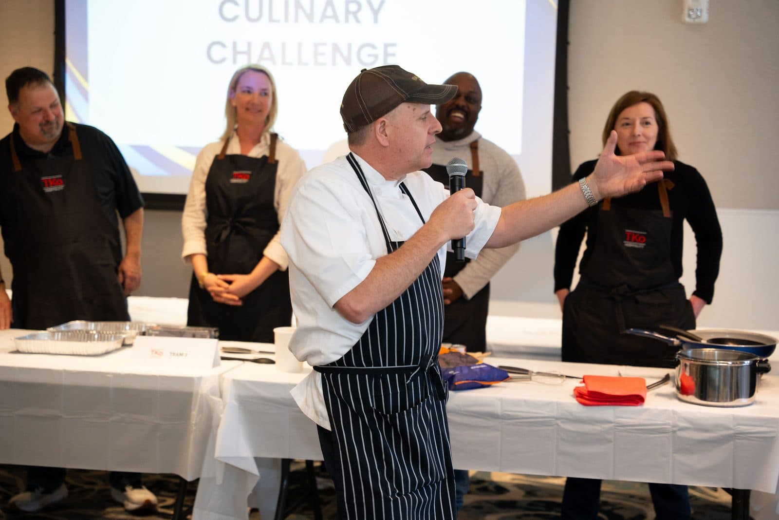 Chef with a microphone leading teams in aprons at a culinary challenge in a ballroom