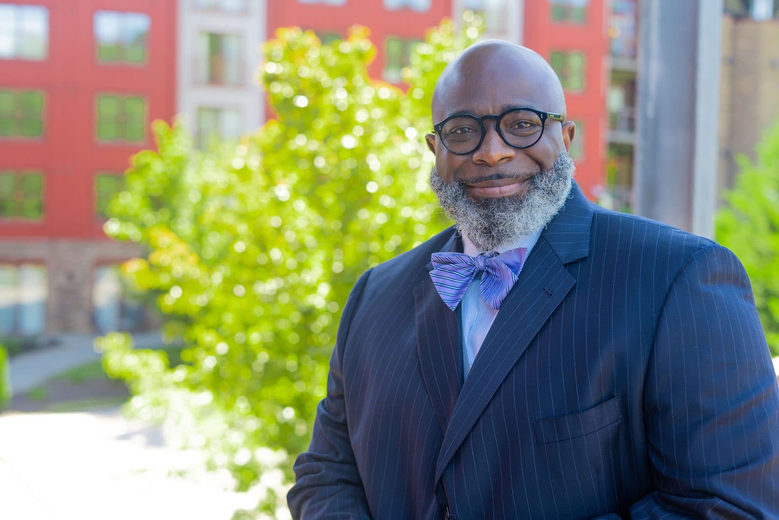 Smiling portrait in a navy pinstripe suit and lavender bow tie outdoors in front of green foliage and a red building