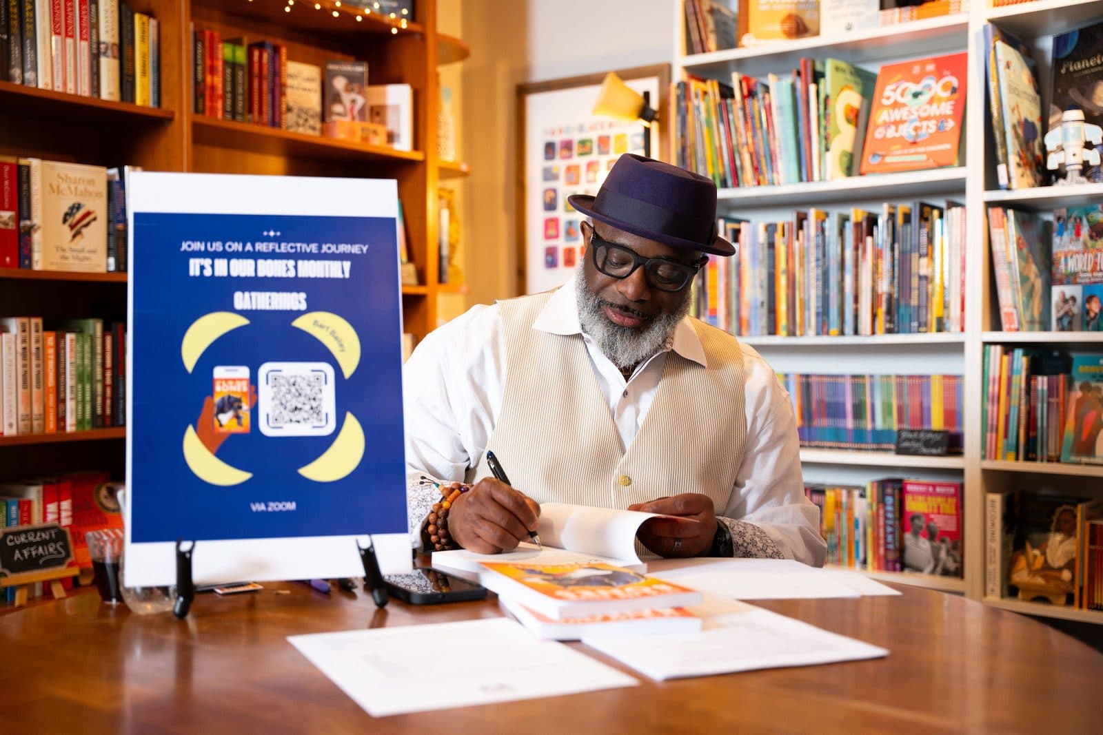 Author in a purple fedora signing a book at a round table in a bookstore, with colorful shelves behind