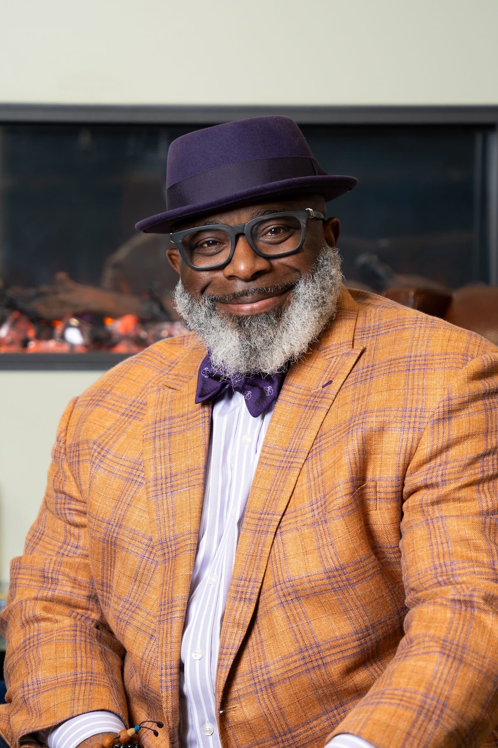 Seated portrait in a purple fedora, glasses, orange plaid blazer and purple bow tie in front of a fireplace