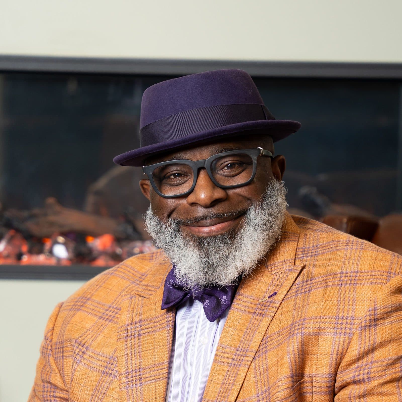 Close-up headshot in a purple fedora, glasses, and orange plaid blazer