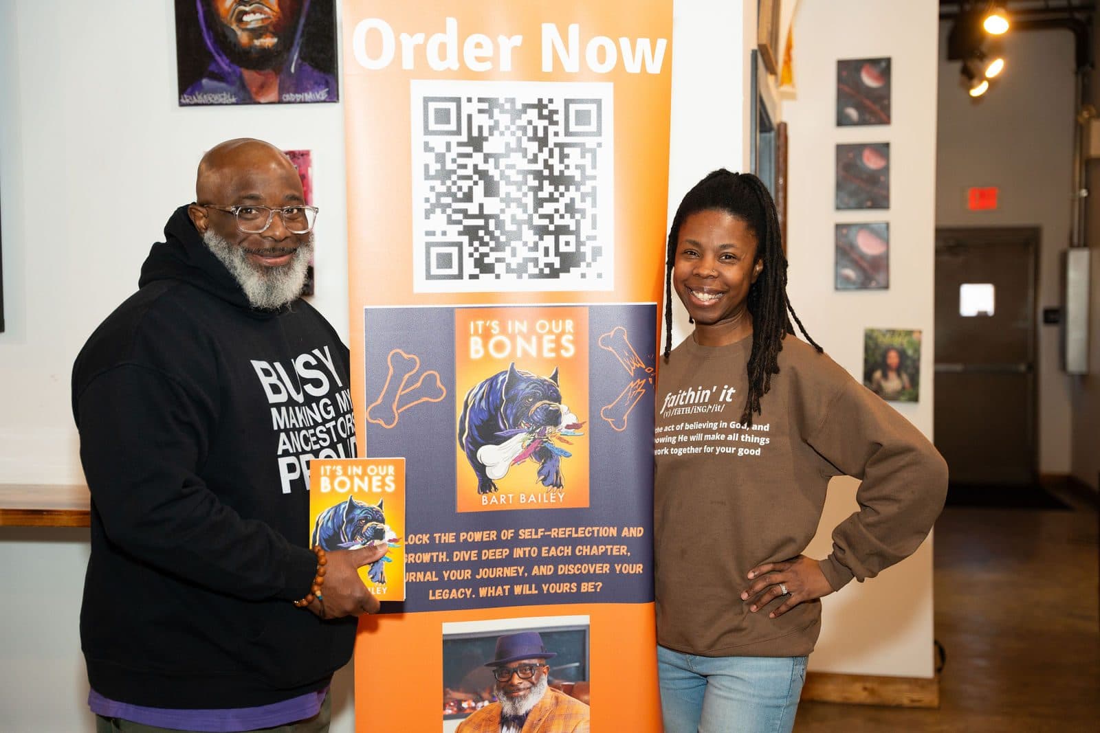 Guest holding the book It's In Our Bones beside another guest, in front of an orange Order Now banner